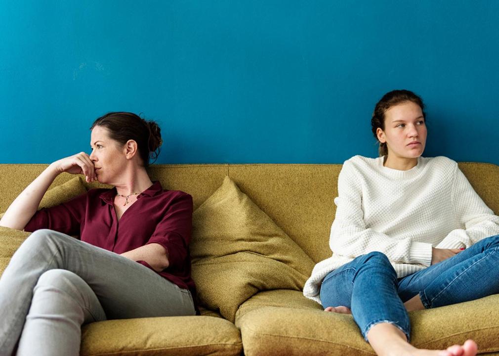 Adult and young adult sitting on opposite sides of yellow couch, looking off into distance in front of blue wall.