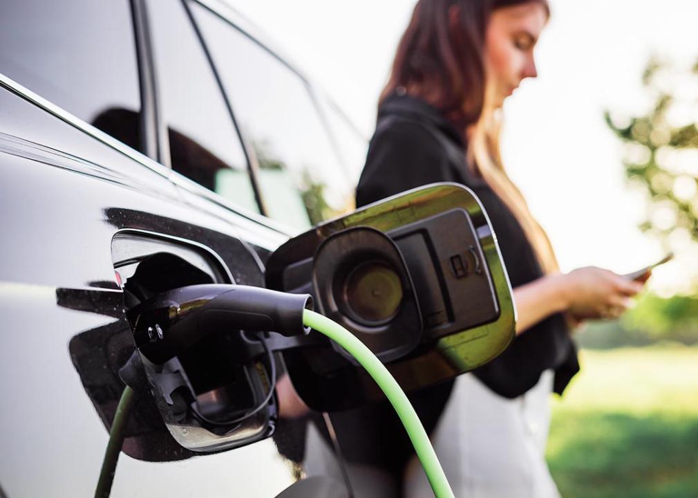 A woman with a phone is leaning on her electric car while it is being charged.