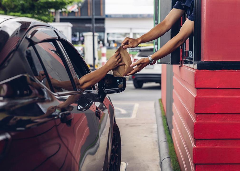 Rear view of drive-thru window worker in red building handing food in brown bag to passenger in passenger seat of black car.