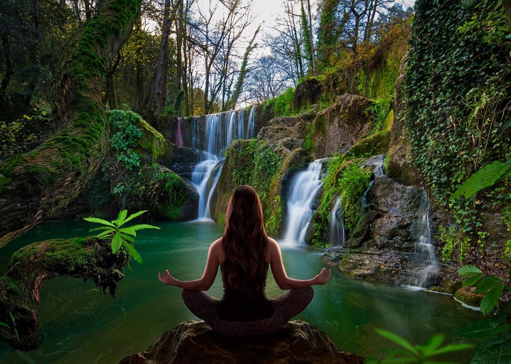 Person sitting in yoga lotus pose facing away from viewer looking at waterfall. 