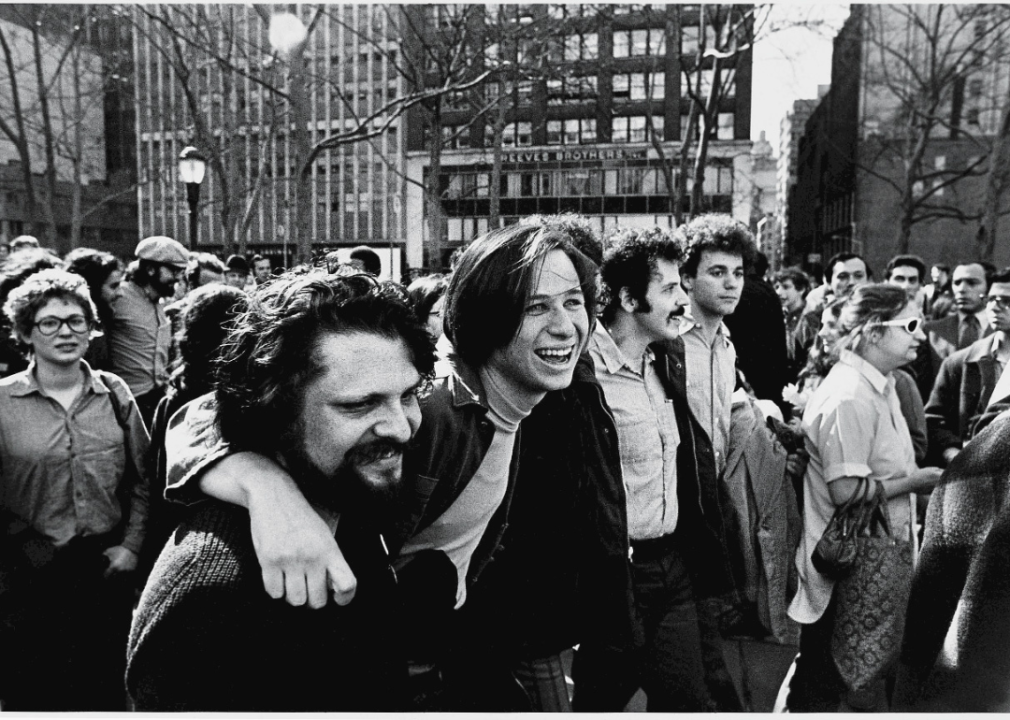 Crowds of protestors in the street, with a focus on a few smiling people with their arms around each other's shoulders. 