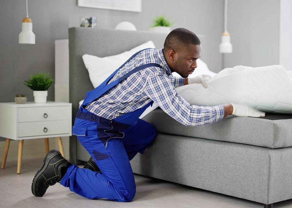 A male exterminator inside a bedroom carefully examining the sheets under a bed.