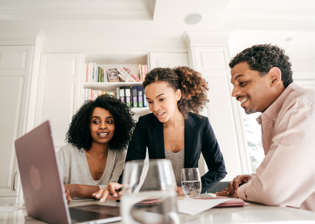 A real estate agent and a couple sat at a table, smiling and looking at a laptop.