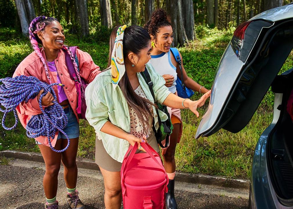Three young people carrying bags for a summer trip load a car trunk.