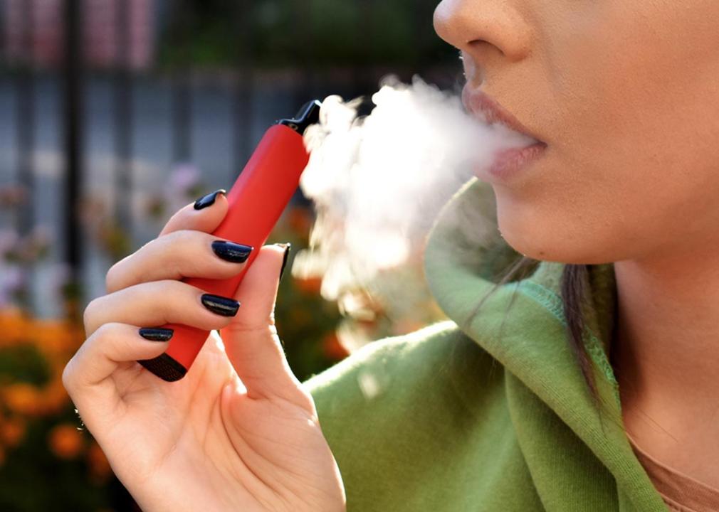 A close up of someone exhaling smoke from vaping, holding vape with black nail polish on fingernails.