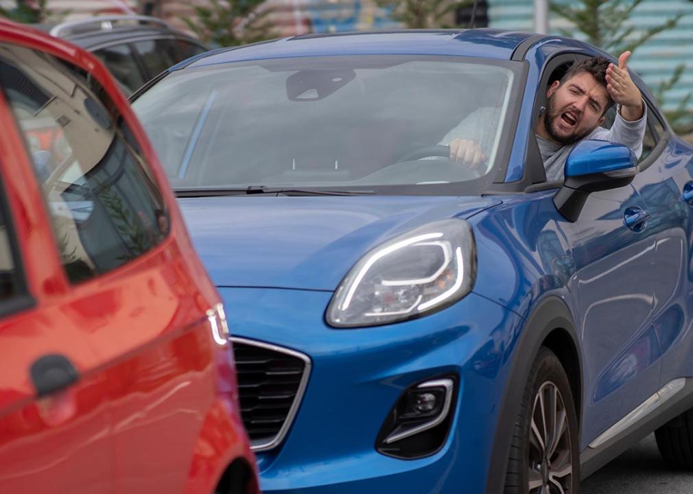 A man driving inside a blue car, his head is sticking out and is seen yelling at a red car in front of him.
