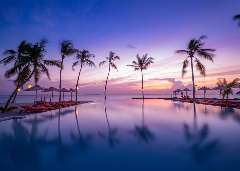 Purple sunset reflected in pool with silhouetted palm trees and ocean in background. 