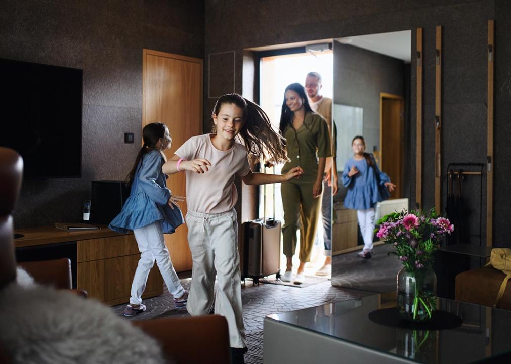 Happy family entering a hotel room with dark decor, parents in doorway with children running in foreground.