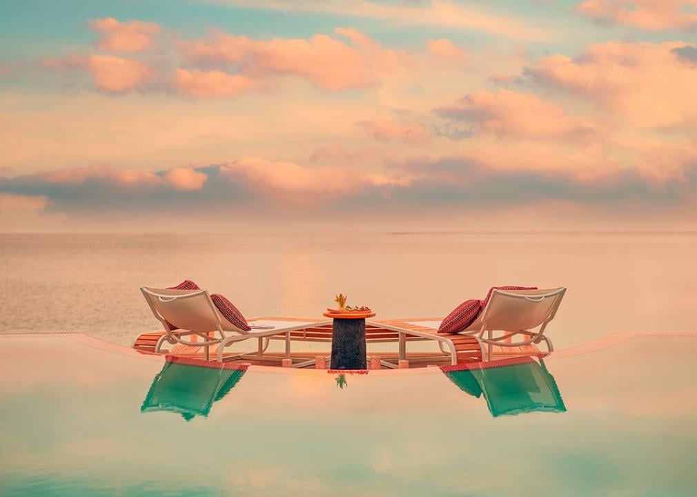 Pink and pastel sunset from the point of view of an infinity pool with two chairs looking out onto the ocean. 