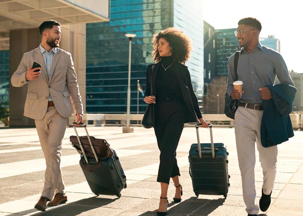 Three professionals in business attire are walking with their suitcases while seemingly having a chat.