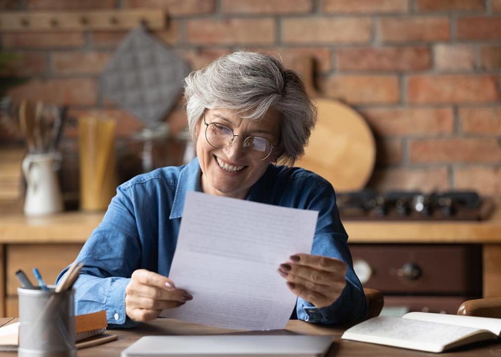 An elderly lady is sitting in her kitchen while happily reading a piece of paper.