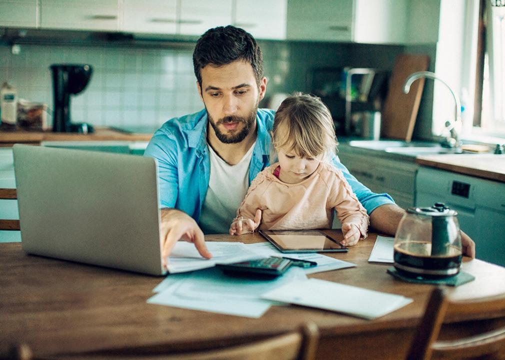 Father sitting at kitchen table with bills, calculator and laptop computer while holding small child.