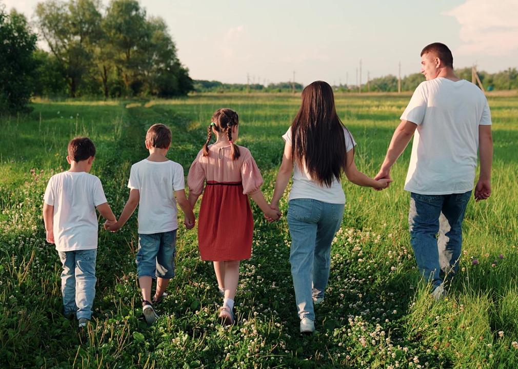 Family of five holding hands seen from the back, walking through a grassy field.
