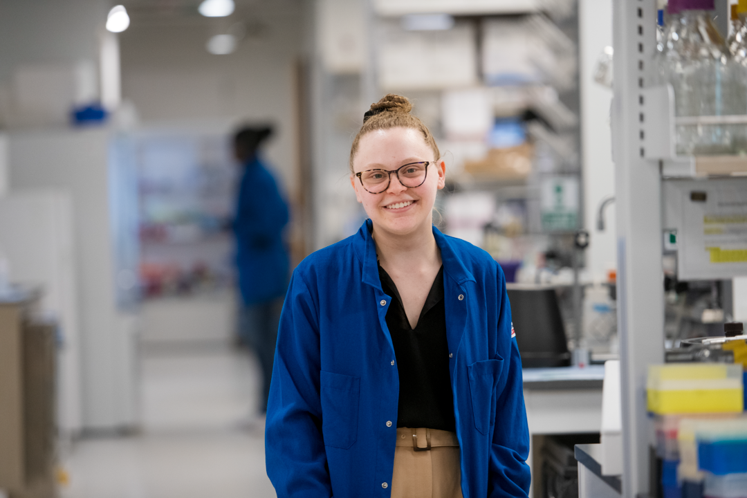 Madeline Szoo wearing a blue labcoat posing for a portrait in a lab. 