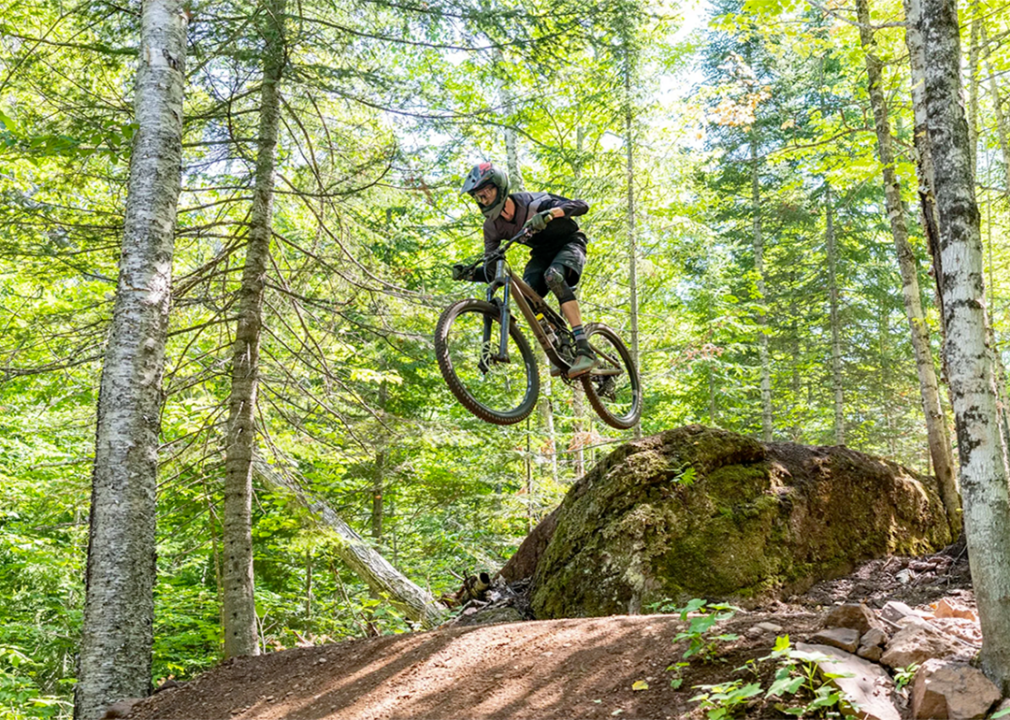 Mountain biker going off a jump on a dirt track with trees in the background. 