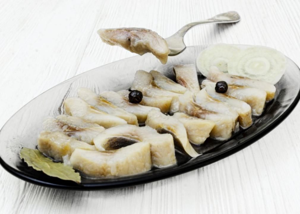 A glass serving dish filled with neatly arranged slices of pickled herring, garnished with onion slices and black peppercorns.