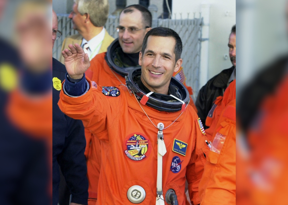 John Herrington in an orange astronaut uniform waving as he walks alongside a couple of other astronauts.
