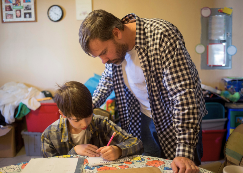 A father stands over young son who is sitting at a table writing on paper.