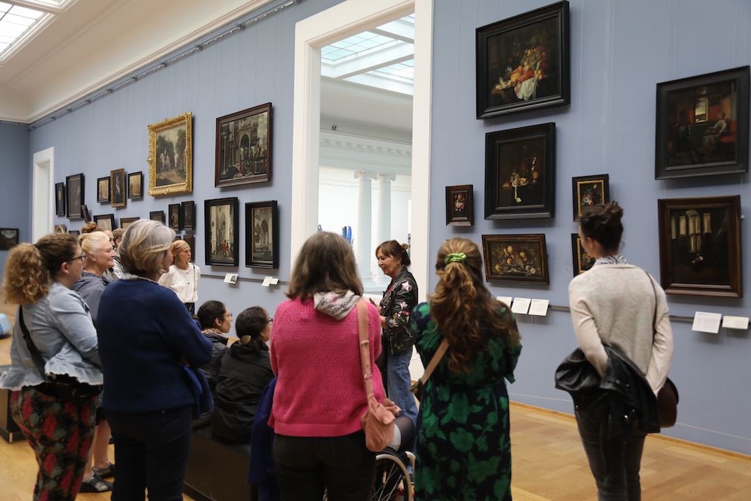 A group on a tour of the museum's collection viewing a room with artwork on pale blue walls. 