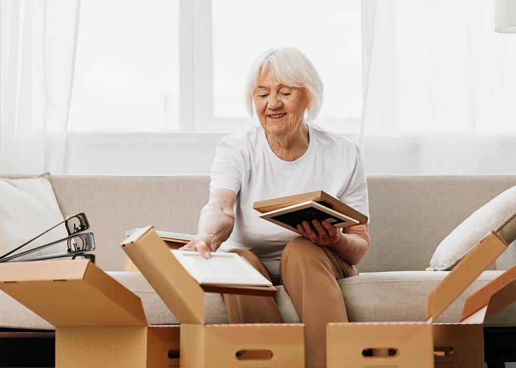 An elderly woman sitting on a sofa collecting home items to put inside boxes.