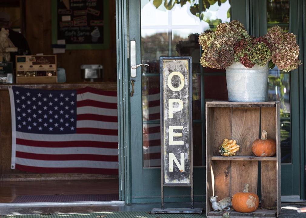 An "open" sign hangs in a shop window with fall decor and American flag in the background. 