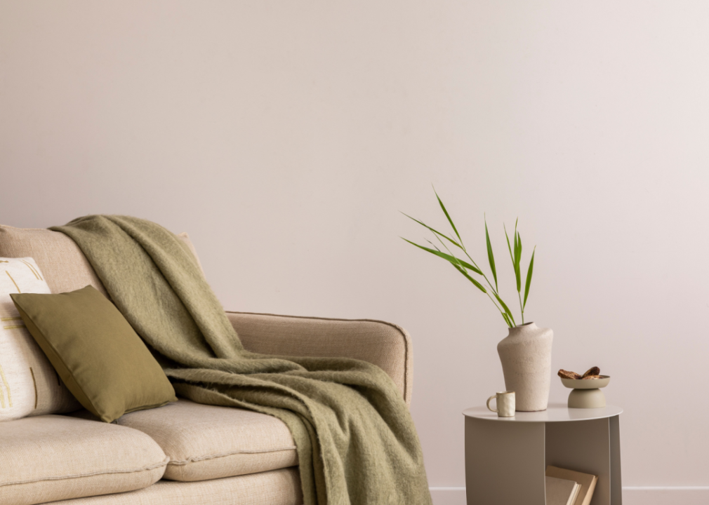 A beige couch with a darker gray blanket is in a minimalist room with a simple table and vase.