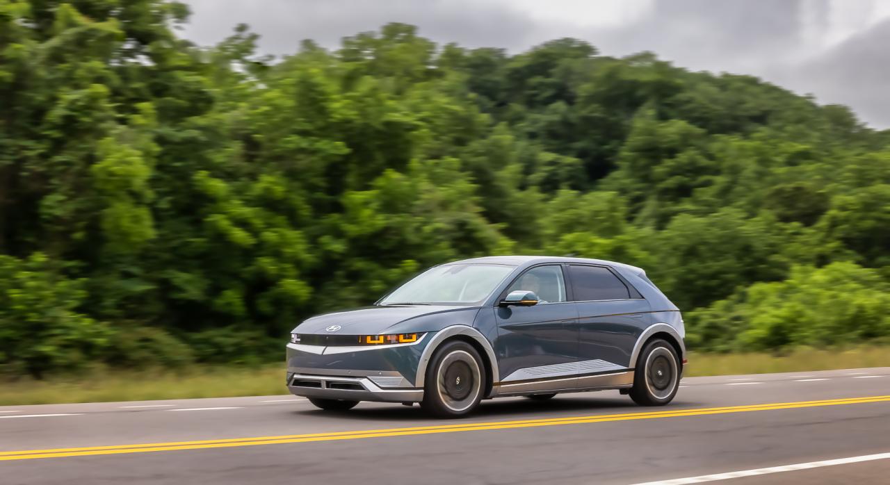 Photo of Dark Hyundai Ioniq 5 driving on road with trees in background. 