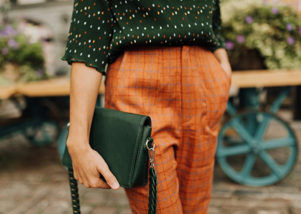 A person wearing slacks and patterned blouse holds a black purse.