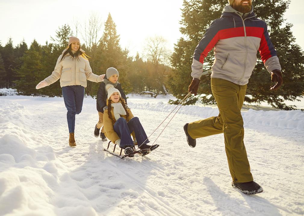 Two parents running outdoors and their two children in sleds during wintertime.