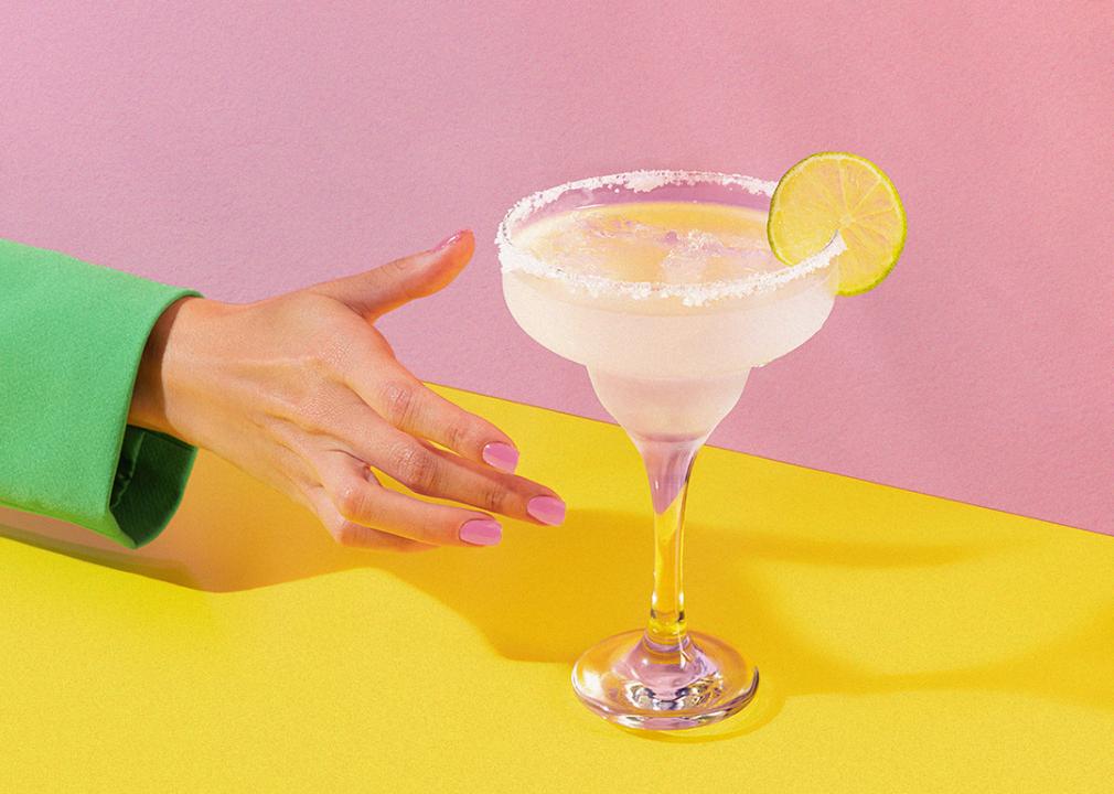 Hand reaching for lemon-garnished drink on a pink and yellow background. 