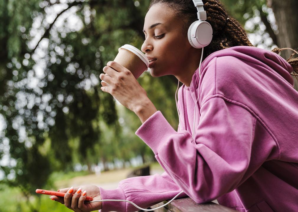 A woman taking a break from working out by drinking coffee while listening to music in the park.