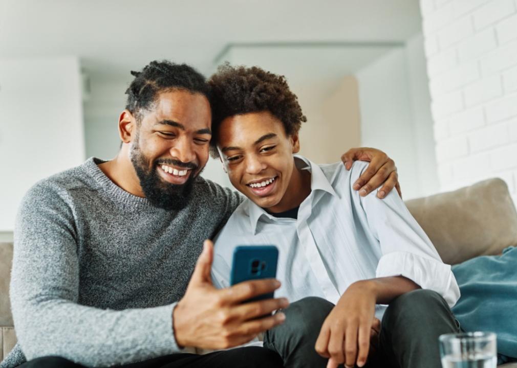 Father and son look at smartphone while sitting on couch.