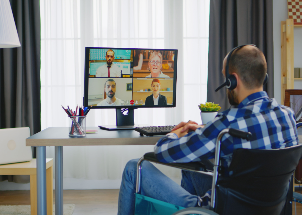 Person wearing a headset in wheelchair videoconferences with colleagues from a remote office.