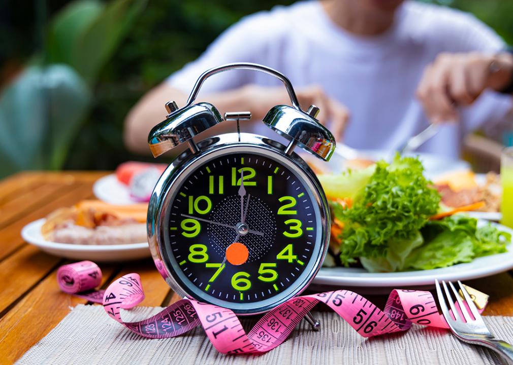 A clock stands in front of a man eating meals, practicing intermittent fasting.