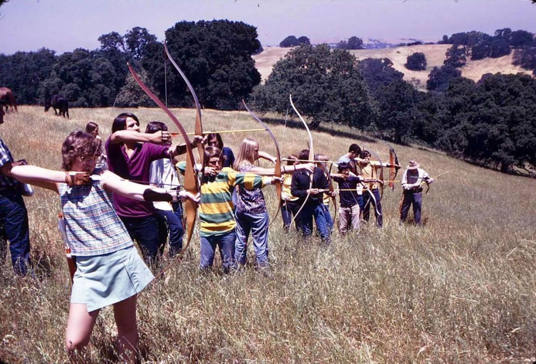 Some teenagers practicing archery in 1973 A line of about a dozen kids all aiming with their archery bows