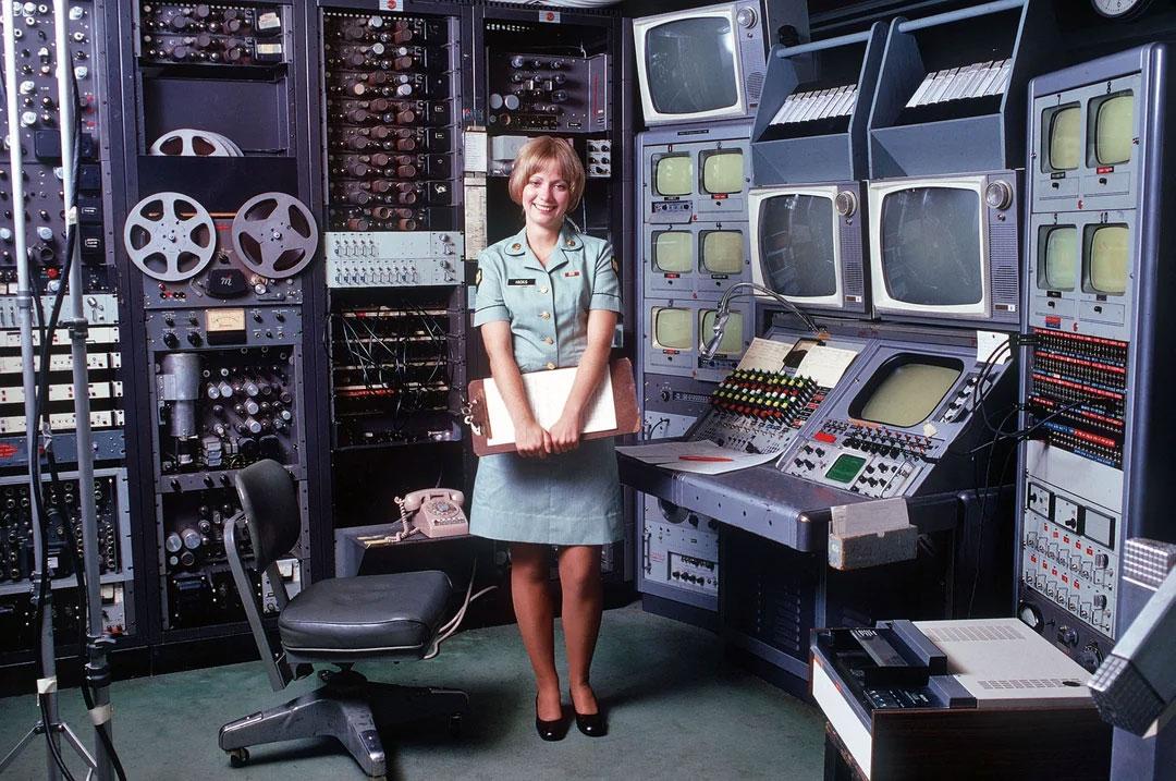 U.S. Army A/V technician stands at her editing station in 1973 A woman in an Army uniform stands in front of a bays of televisions and audio devices