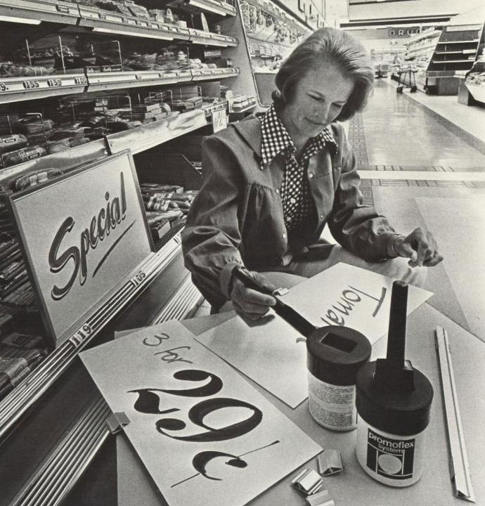 A woman painting a Supermarket sign in the aisle of a grocery store Woman sitting at a table in a grocery store painting the sale signs