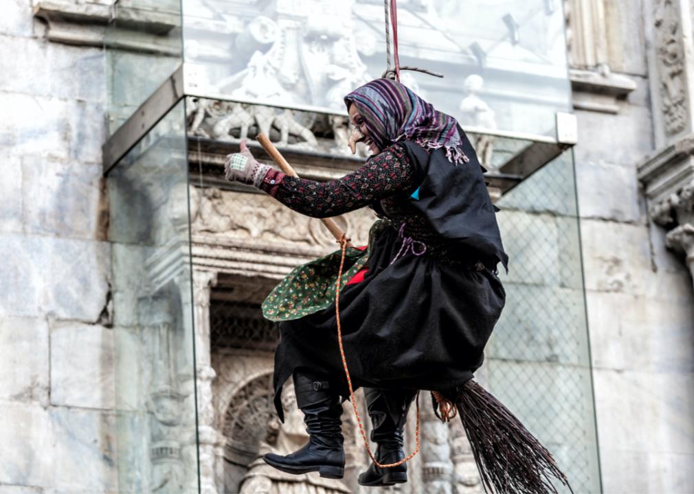 A person dressed as a witch riding a broom and suspended in the air by ropes.