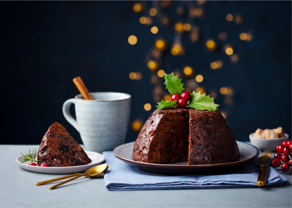 A Christmas pudding with holly and berries on top of it next to a slice of the dessert and a mug with a cinnamon stick in it.