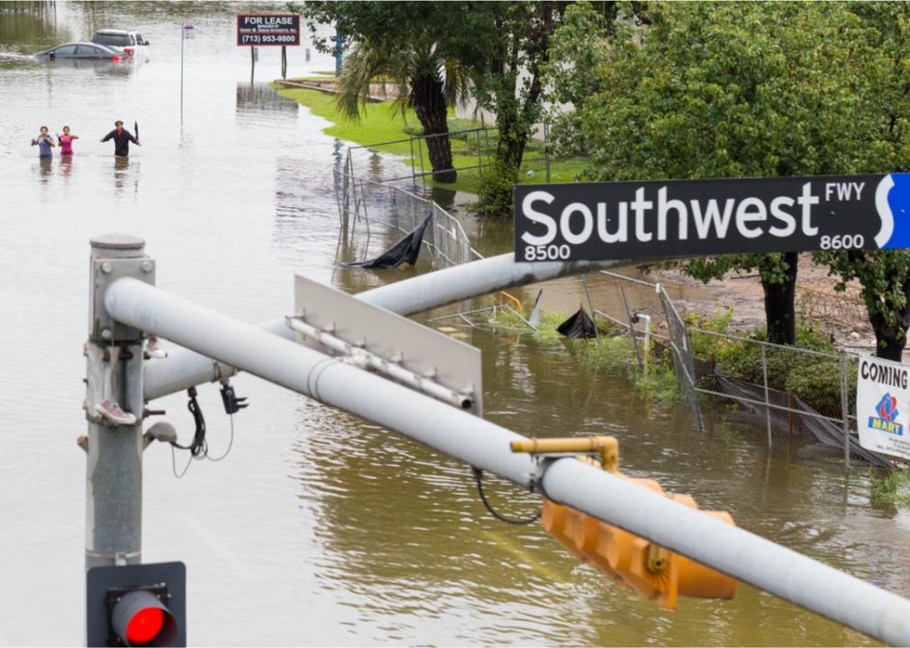 Photo shows a flooded neighborhood with water halfway up a pole that holds a stoplight and people wading in waist-deep water