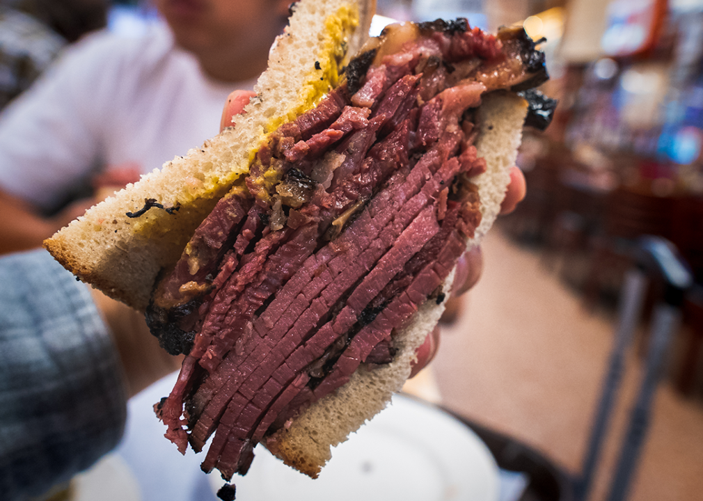 Hand holding thick cut pastrami sandwich on rye with mustard.