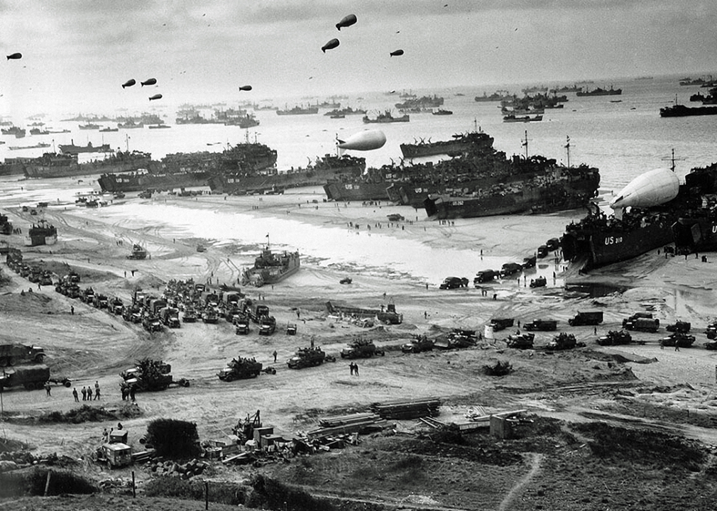 A large number of ships in front of Omaha Beach delivering troops, equipment and vehicles lining up to leave the beach, barrage balloons are up in the sky.