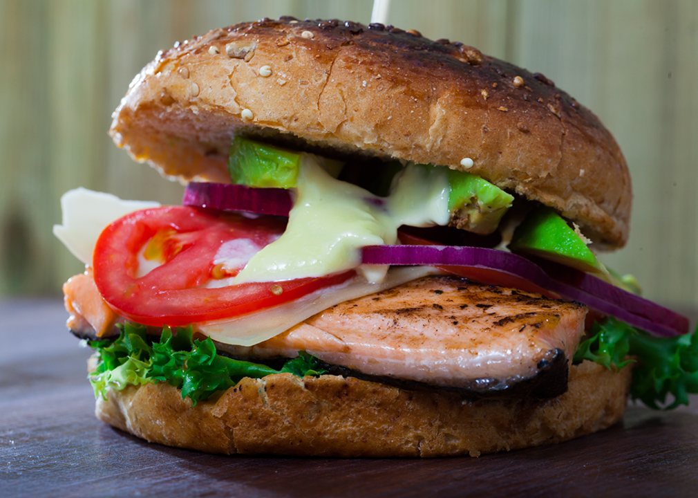 Close up of a roasted trout, avocado, lettuce, tomato on roll.