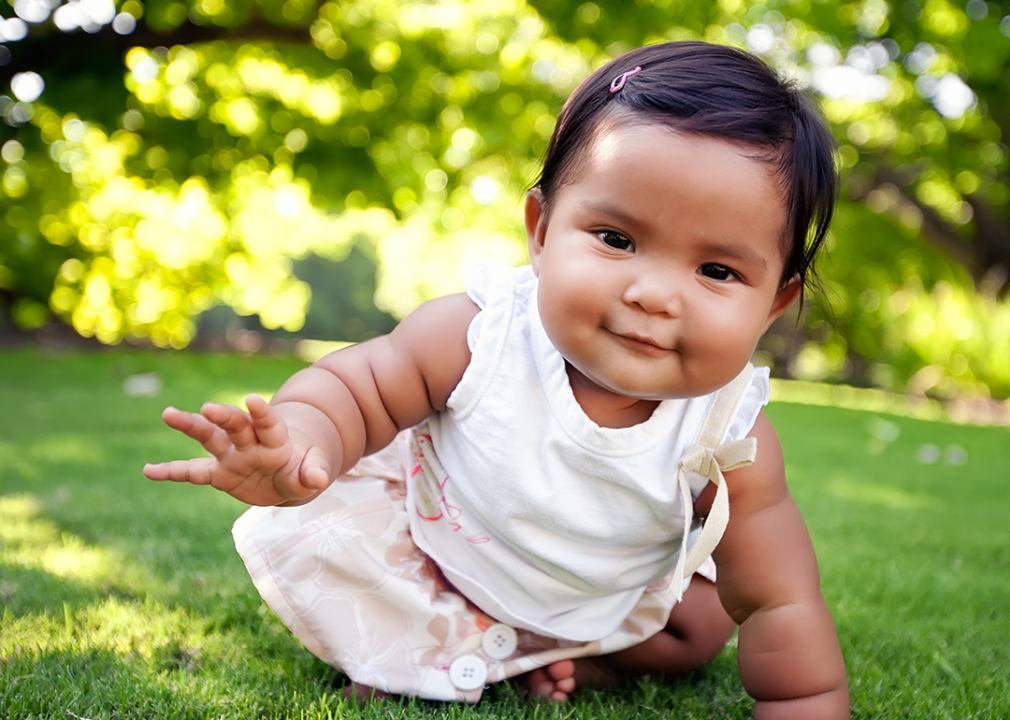 Baby sitting on grass reaching out to camera.