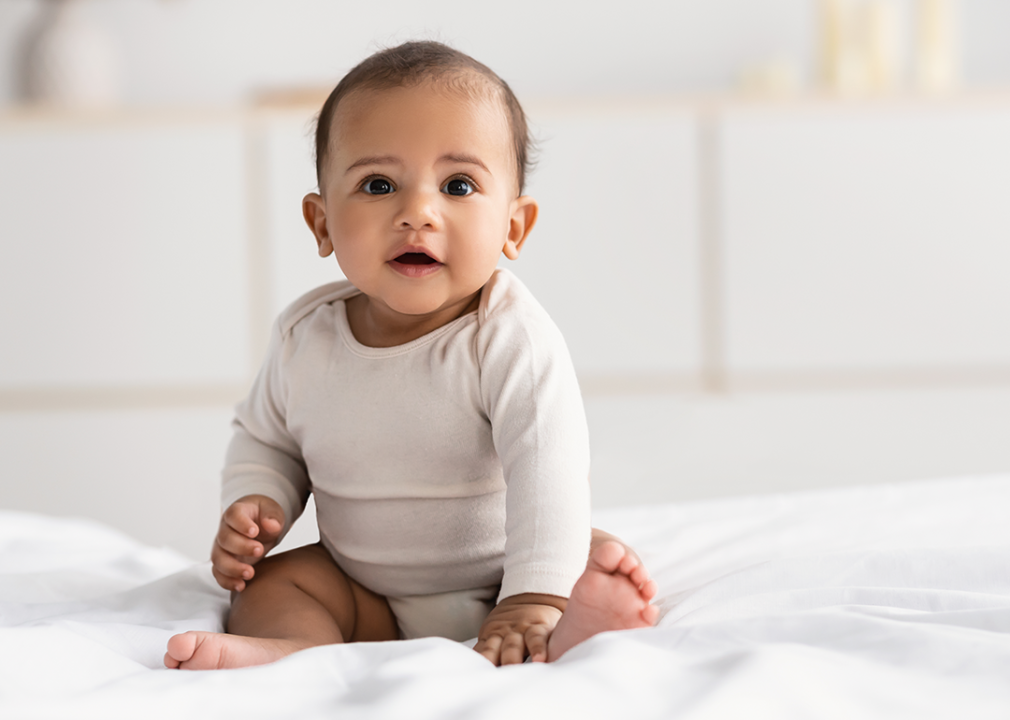 Baby wearing onesie sitting on bed.