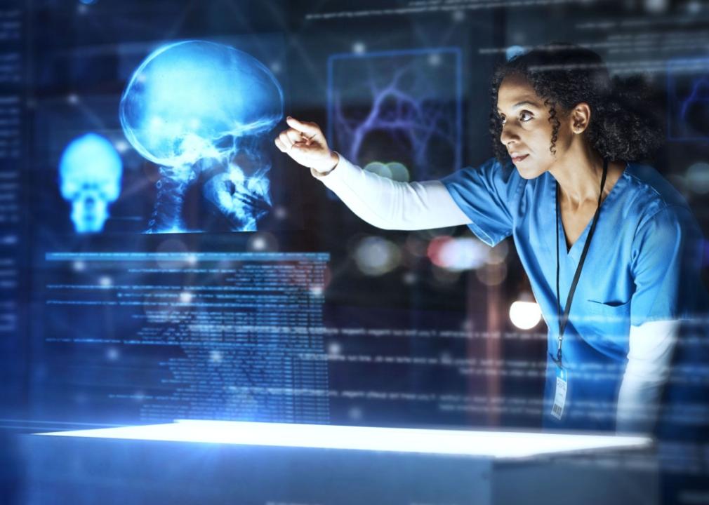 A woman in scrubs is standing in a dark room pointing at the large hologram of a skull. 