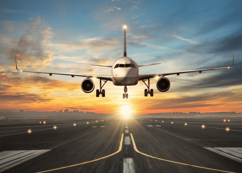 An airplane during its final approach for landing on a runway. Bright orange sun positioned low on the horizon. 