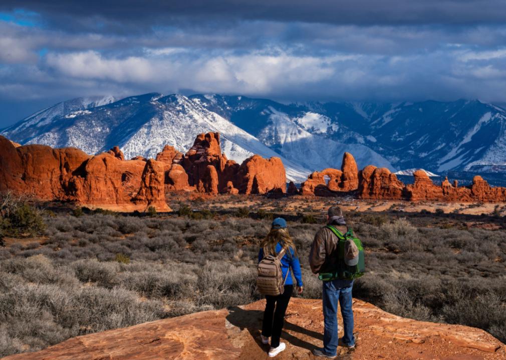 A couple looking at bright orange rock formations with snowy taller mountains in the distance.