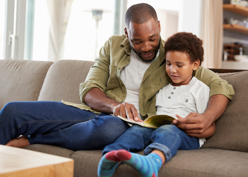A Black father reading to his son on a couch.