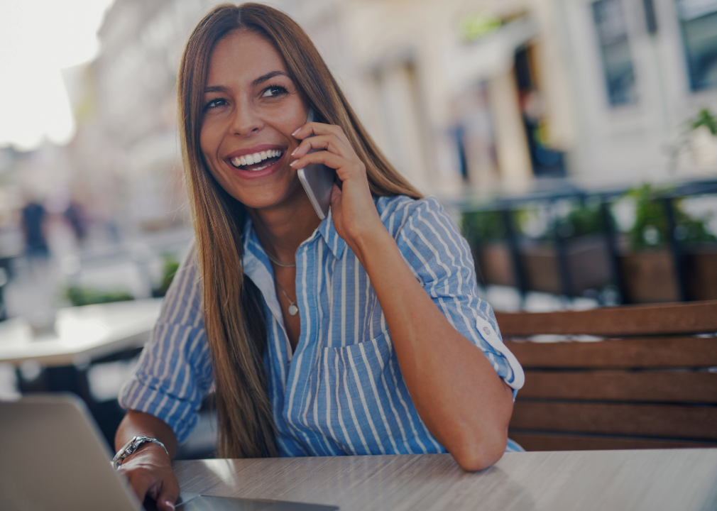 A young woman with long hair and a big smile sitting at the table outdoors holding a cellphone to her ear.